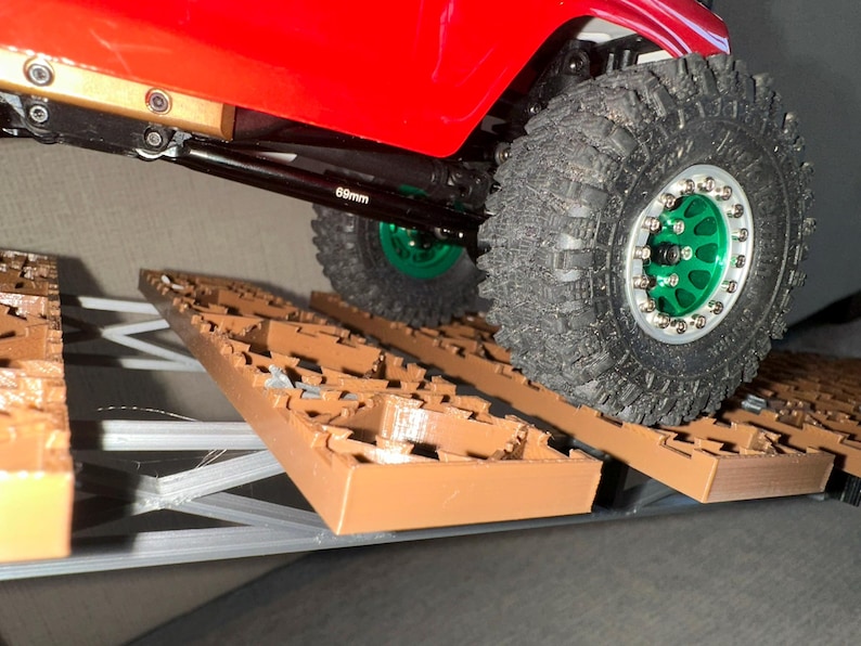 May include: A red toy truck with black axles and green wheels is driving over a brown Lego-like obstacle course. The truck has a 60mm axle.