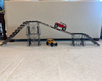 May include: A red and a yellow toy truck driving up a ramp made of gray metal and brown plastic. The ramp is made of multiple sections that can be connected together.