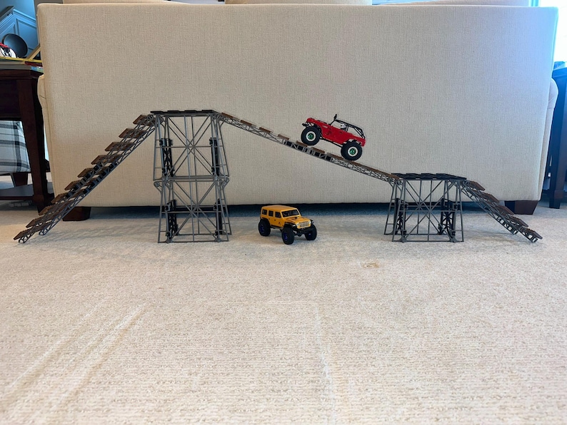 May include: A red and a yellow toy truck driving up a ramp made of grey metal and brown plastic. The ramp is made of multiple sections that can be connected together.