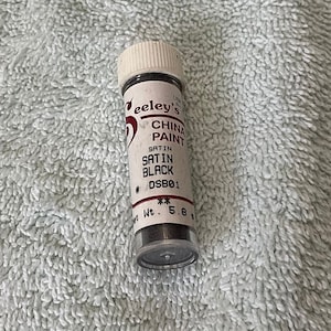 Puede incluir: Un pequeño frasco de pintura para porcelana Seeley's en negro satinado, etiquetado como DSB01. El frasco es blanco con letras negras y una tapa negra. El frasco contiene una pequeña cantidad de pintura negra.