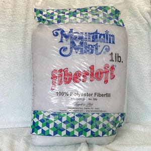 Puede incluir: Una bolsa de plástico blanca con un patrón geométrico azul y verde en la parte inferior. La bolsa contiene fibra de relleno blanca y tiene una etiqueta que dice "Mountain Mist fiberloft 1 lb. 100% Polyester Fiberfill 1 lb. (454 g) No. 300 Leggett & Platt 3307 Walden Ave., Depew, NY 14043 mountainmeggett.com www.mountainmisttp.com".