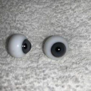 May include: Two spherical doll eyes with white sclera and dark grey irises featuring a small black pupil in the centre. The eyes are smooth and rounded, designed for use in crafting or doll making, and are presented as a pair of matching components.