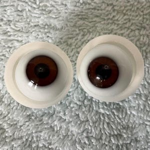 Peut inclure: Deux yeux de poupée en verre brun avec une pupille noire et un iris blanc, placés dans des orbites en plastique blanc.