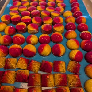 Könnte beinhalten: Draufsicht auf ein blaues Tablett mit bunten Pralinen. Die Pralinen sind rund und pyramidenförmig, mit Farbverläufen von Rot und Gelb. Die Pralinen sind in ordentlichen Reihen angeordnet und bilden eine ansprechende Präsentation.