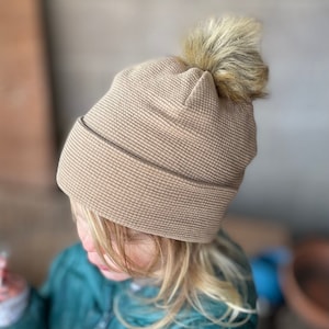May include: A tan waffle knit beanie with a brown faux fur pom pom on top.
