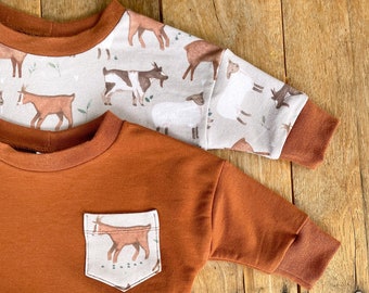 May include: A rust-colored long-sleeved sweatshirt with a pocket featuring a brown goat illustration. The sweatshirt has a ribbed hem and cuffs. The sweatshirt is on a wooden surface with a white circle sticker that says "spotted fawn Handmade Cloth Diapers & More".