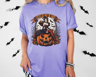 May include: A light purple t-shirt featuring a Halloween graphic. The graphic includes a witch sitting on a jack-o'-lantern with the text "SEASON OF THE WITCH" on a banner. The background includes a full moon, bats, and trees.
