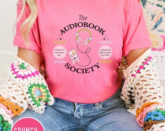 May include: Pink t-shirt with a graphic design of a phone with headphones and the text "The Audiobook Society". The text "Oh that's how to pronounce that" and "Multitasking in different worlds since 1990s" are also included in the design.