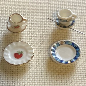 May include: Four miniature ceramic tea sets, each consisting of a small cup and saucer. The designs include black and white floral patterns, a red strawberry motif, a blue and white geometric border, and a blue floral pattern with a metal needle.
