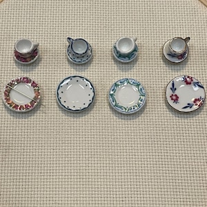 May include: A set of four miniature ceramic teacups and four matching saucers arranged in two rows. The pieces feature various patterns including pink floral, blue polka dots, green scrollwork, and red floral designs. A thin metal needle lies across one saucer.