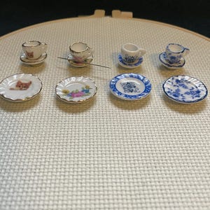 May include: Miniature porcelain tea sets with saucers. One set features a cat design, while others have floral or blue patterns. The tiny cups and saucers are arranged on a white surface, perfect for dollhouses or collectors.
