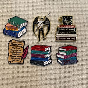 May include: Collection of enamel pins featuring book stacks, a winged figure, and a teacup. The pins have vibrant colors and include text such as "A Court of Thorns and Roses" and "Rhysand is the most handsome High Lord."