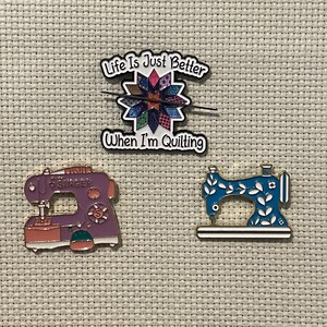 May include: Three sewing-themed enamel pins. One pin reads "Life Is Just Better When I'm Quilting" above a colorful quilt design. The other two pins are sewing machine designs, one purple and one blue.