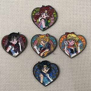 May include: Five heart-shaped enamel pins featuring anime characters. Each pin has a different character with colorful details and a stained-glass style border. The pins are set against a neutral, textured background.
