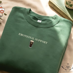 May include: A dark green sweatshirt with the words "EMOTIONAL SUPPORT" printed in white. Below the text is a small graphic of a pint of stout. The sweatshirt is made of a soft material.