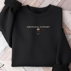 May include: Black sweatshirt with the words "EMOTIONAL SUPPORT" above a graphic of a pint of stout. The sweatshirt has a crew neck and long sleeves. The design is centered on the chest.