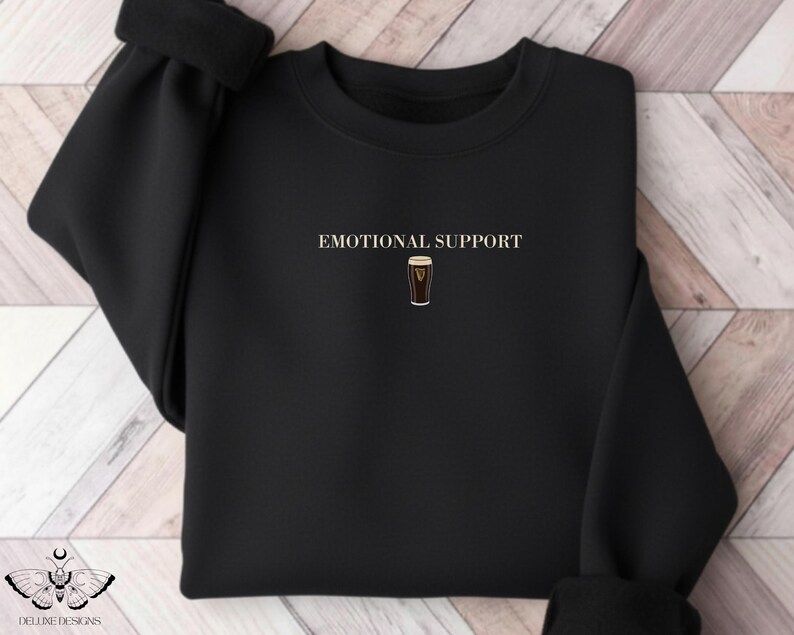 May include: Black sweatshirt with the words "EMOTIONAL SUPPORT" above a graphic of a pint of stout. The sweatshirt has a crew neck and long sleeves. The design is centered on the chest.