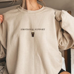 May include: Beige sweatshirt with the text "EMOTIONAL SUPPORT" above a graphic of a pint of stout. The sweatshirt has long sleeves and a crew neck. The design is centered on the chest.