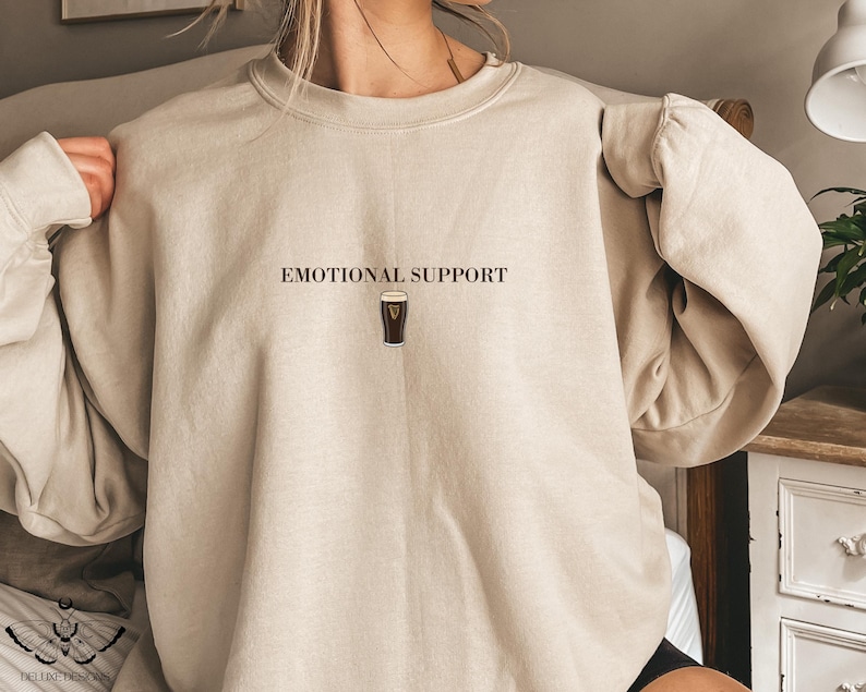 May include: Beige sweatshirt with the text "EMOTIONAL SUPPORT" above a graphic of a pint of stout. The sweatshirt has long sleeves and a crew neck. The design is centered on the chest.
