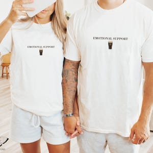 May include: Two off-white t-shirts with the text "EMOTIONAL SUPPORT" above a graphic of a pint of stout. The shirts are worn by a couple, one wearing gray shorts. The image is taken in a well-lit room.