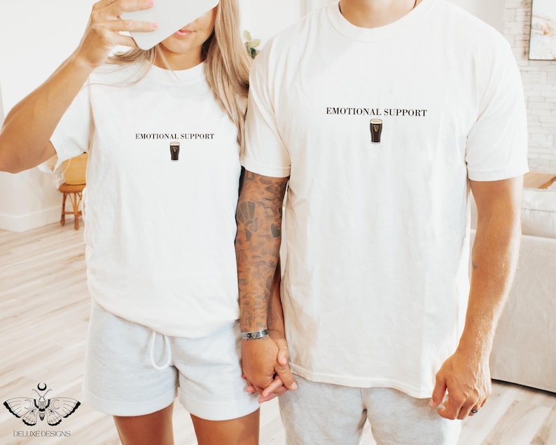 May include: Two off-white t-shirts with the text "EMOTIONAL SUPPORT" above a graphic of a pint of stout. The shirts are worn by a couple, one wearing gray shorts. The image is taken in a well-lit room.