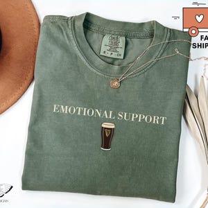 May include: A sage green t-shirt with the words "EMOTIONAL SUPPORT" printed in a serif font, along with an illustration of a pint of stout. A gold necklace rests on the shirt. The shirt is folded and displayed with a hat and dried plant.