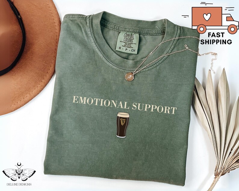 May include: A sage green t-shirt with the words "EMOTIONAL SUPPORT" printed in a serif font, along with an illustration of a pint of stout. A gold necklace rests on the shirt. The shirt is folded and displayed with a hat and dried plant.