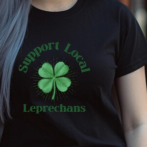 May include: A black short-sleeved t-shirt featuring a green four-leaf clover graphic centered on the chest. The text "Support Local" is arched above the clover, and the word "Leprechans" is printed in green below the clover design.