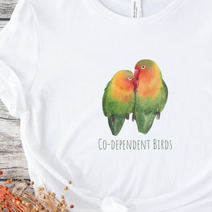 May include: White t-shirt featuring a watercolor illustration of two colorful birds, with green, orange, and red plumage, nestled together. The text "CO-DEPENDENT BIRDS" is printed below the birds. The shirt is made of a soft material.