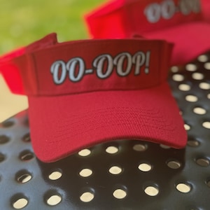 May include: Red visor with the text "OO-OOP!" in white, with a curved brim. The visor is made of a textured fabric. Other visors are visible in the background, out of focus.