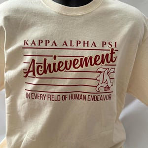 May include: Cream-colored t-shirt with maroon text. The text reads "KAPPA ALPHA PSI Achievement" with a stylized "K" and the year "1911". Below, it says "IN EVERY FIELD OF HUMAN ENDEAVOR".