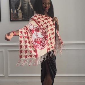 May include: A cream-colored shawl with a red houndstooth pattern and fringe. The shawl features a large emblem with Greek letters and text. The shawl is draped over the shoulders.
