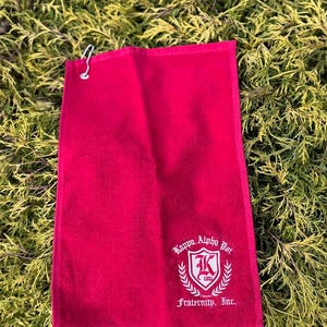 May include: A red towel with a metal hook, featuring the text "Kappa Alpha Psi Fraternity, Inc." in white. The towel has a shield design with the letter "K" and the year 1911. The towel is lying on a bed of green foliage.