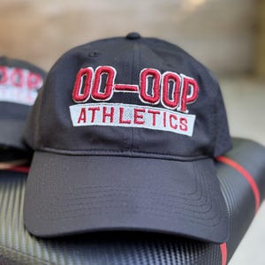 May include: Black baseball cap with the text "00-OOP ATHLETICS" embroidered in red and white. The cap has a curved brim and a button on top. The hat is sitting on a textured surface.