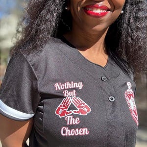 May include: Black baseball jersey with white sleeve stripes. The jersey features the text "Nothing But The Chosen" in red and white, along with a red hand symbol. A crest is visible on the right side.