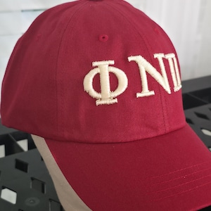 May include: A maroon baseball cap with the Greek letters ΦNI embroidered in off-white. The cap has a curved brim and a button on top. The hat is displayed on a mannequin head.