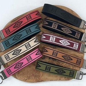 May include: Eight colorful leather keychains with a geometric pattern and silver key rings. The keychains are arranged on a wooden surface.