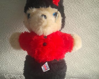 May include: A plush toy bear dressed as a British Royal Guard. The bear has a black hat with a red accent, a red jacket, and black pants. The bear has a tag with the Union Jack flag.