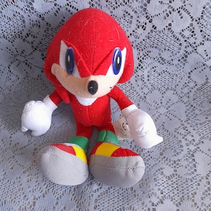 May include: A plush toy of the character Knuckles from the Sonic the Hedgehog series. The figure features bright red fur, large white gloves, and shoes coloured in red, yellow, and grey. It has blue eyes and a small black nose, with a white tag attached.