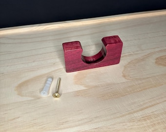May include: A small, handcrafted, deep red wooden bracket with a curved groove, designed to hold an item. Includes a white plastic wall anchor and a gold-colored screw for mounting. The bracket is made of solid wood and has a smooth finish.