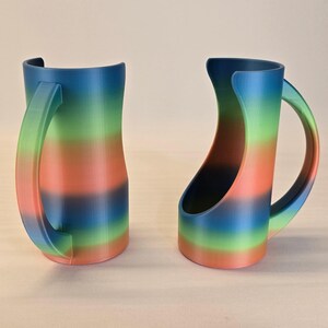 May include: Two rainbow-colored mugs with curved handles. The mugs feature a gradient of colors, including blue, green, orange, and red. The handles are a lighter shade of the same colors. The mugs are designed with a unique, open-sided shape.