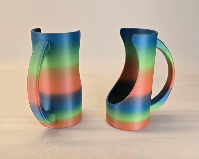 May include: Two rainbow-colored mugs with curved handles. The mugs feature a gradient of colors, including blue, green, orange, and red. The handles are a lighter shade of the same colors. The mugs are designed with a unique, open-sided shape.