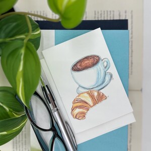 May include: A white coffee cup with brown coffee inside, a croissant, and a silver pen on top of a stack of books. The top book is light blue, the middle book is dark blue, and the bottom book is white.