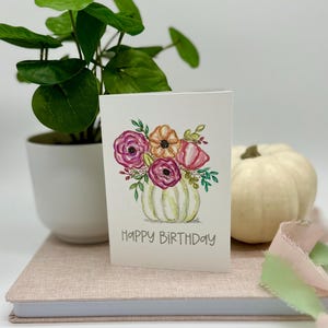 May include: A white card with a watercolor illustration of a white pumpkin vase with pink, orange, and red flowers. The text "Happy Birthday" is printed below the vase.