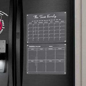 May include: Black fridge with a water dispenser and ice maker. Two clear acrylic magnetic boards are attached, one with a calendar and the other with a weekly plan. The text "The Tocus Family" is at the top of the calendar board. Includes 3 free markers.