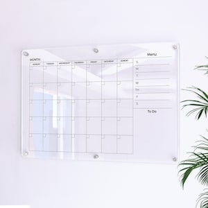 May include: A clear acrylic dry-erase calendar and to-do list board with a monthly calendar grid, a menu section, and a to-do list section. The board is mounted on a white wall.