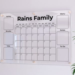 May include: A clear acrylic dry-erase calendar with the text "Rains Family" at the top. The calendar has spaces for each month of the year, as well as sections for notes, to do list, and individual names: Ashley, Brooke, and Talia.