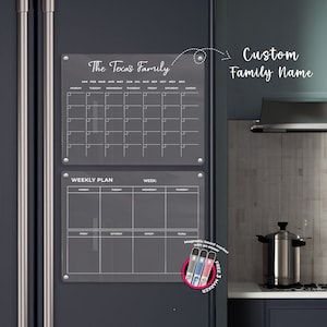 Könnte beinhalten: Transparenter Familienkalender und Wochenplaner aus Acryl, befestigt an einem dunkelgrauen Kühlschrank. Der Kalender trägt die Aufschrift "The Texas Family" und enthält ein monatliches Layout. Der Wochenplaner befindet sich darunter. Enthält ein Set magnetischer Boardmarker.