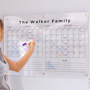 Pode incluir: Um quadro de calendário de parede em acrílico transparente intitulado The Walker Family, apresentando duas grelhas mensais para outubro e novembro com notas manuscritas em várias cores, uma secção de tarefas e quatro suportes de montagem prateados.