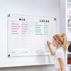 May include: A clear acrylic wall-mounted chore chart with two columns labelled MIA and LUCAS. Each section features a list of chores, checkboxes for days of the week, and fields for points and earnings. The board is filled with handwritten text.
